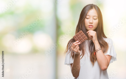 Young asian woman eating chocolate bar over isolated background serious face thinking about question  very confused idea