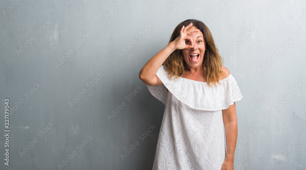 Middle age hispanic woman standing over grey grunge wall doing ok gesture with hand smiling, eye looking through fingers with happy face. Middle age hispanic woman standing over grey grunge wall doing ok gesture with hand smiling, eye looking through fingers with happy face.
