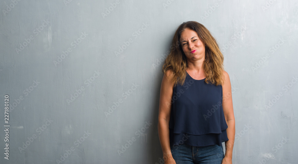 Middle age hispanic woman standing over grey grunge wall puffing cheeks with funny face. Mouth inflated with air, crazy expression. Middle age hispanic woman standing over grey grunge wall puffing cheeks with funny face. Mouth inflated with air, crazy expression.