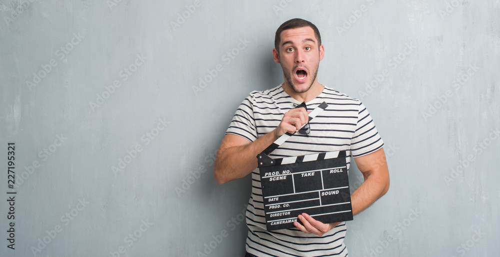 Young caucasian man over grey grunge wall holding movie clapboard scared in shock with a surprise face, afraid and excited with fear expression Young caucasian man over grey grunge wall holding movie clapboard scared in shock with a surprise face, afraid and excited with fear expression