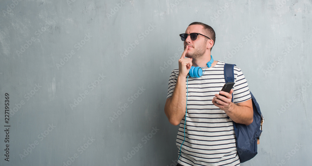 Young caucasian student man over grey grunge wall wearing headphones using smartphone serious face thinking about question, very confused idea