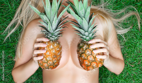 A young woman holds pineapples near her breast while lying on the grass. Bra from pineapples. Tropical modesty.
