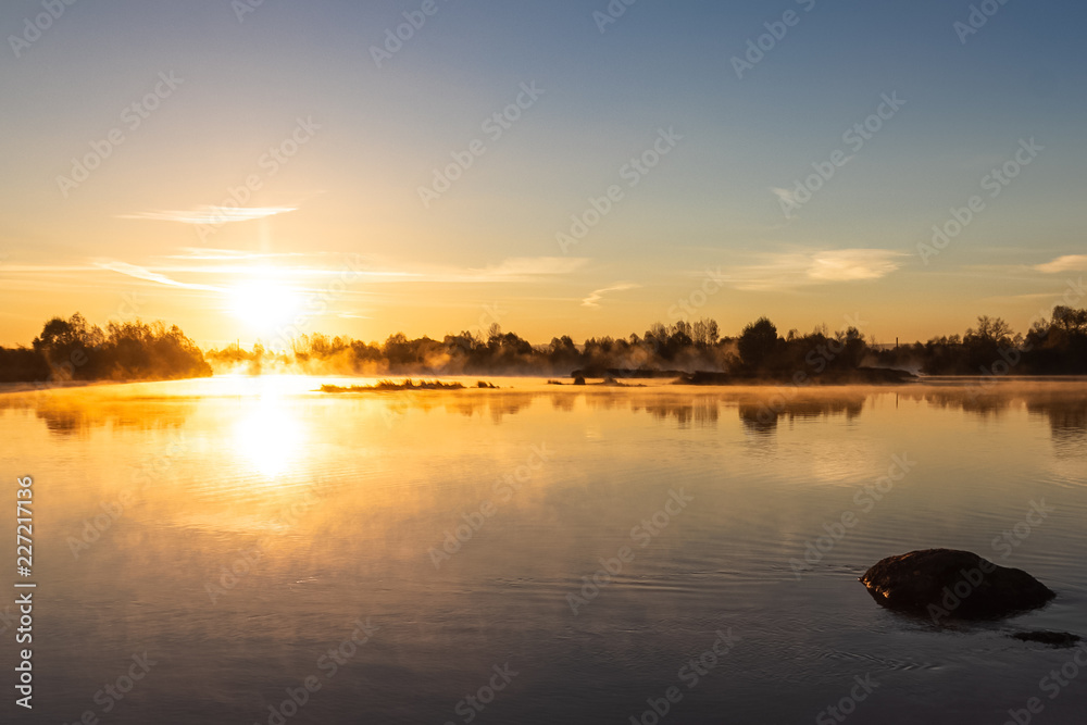 Obraz premium Bright orange dawn warm rays dispel mist over a calm river, illuminating the light in the area.