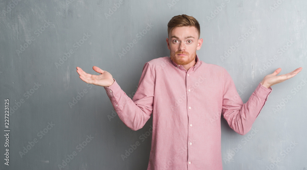 Young redhead man over grey grunge wall wearing pink shirt clueless and confused expression with arms and hands raised. Doubt concept. Young redhead man over grey grunge wall wearing pink shirt clueless and confused expression with arms and hands raised. Doubt concept.