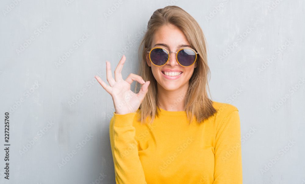 Fototapeta premium Piękna młoda kobieta stojąc na szarej ścianie grunge w okularach retro robi ok znak palcami, doskonały symbol