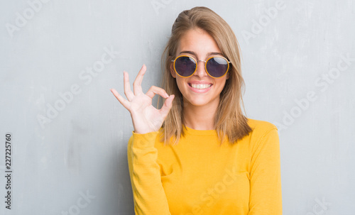 Beautiful young woman standing over grunge grey wall wearing retro sunglasses doing ok sign with fingers, excellent symbol