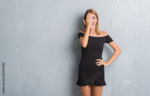 Beautiful young woman standing over grunge grey wall wearing elegant dress covering one eye with hand with confident smile on face and surprise emotion. © Krakenimages.com
