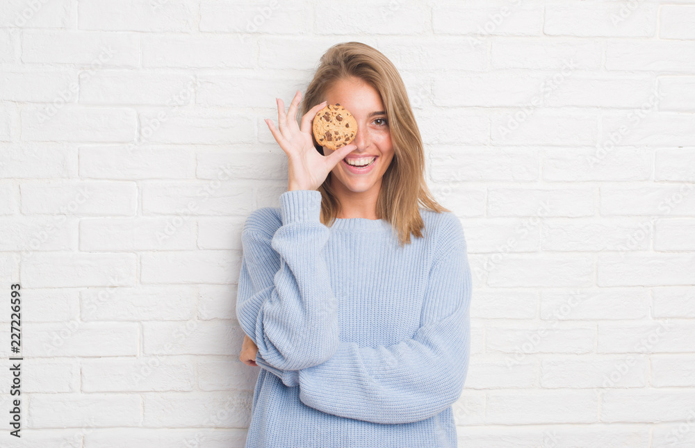 Beautiful young woman over white brick wall eating chocolate chip cooky with a happy face standing and smiling with a confident smile showing teeth Beautiful young woman over white brick wall eating chocolate chip cooky with a happy face standing and smiling with a confident smile showing teeth