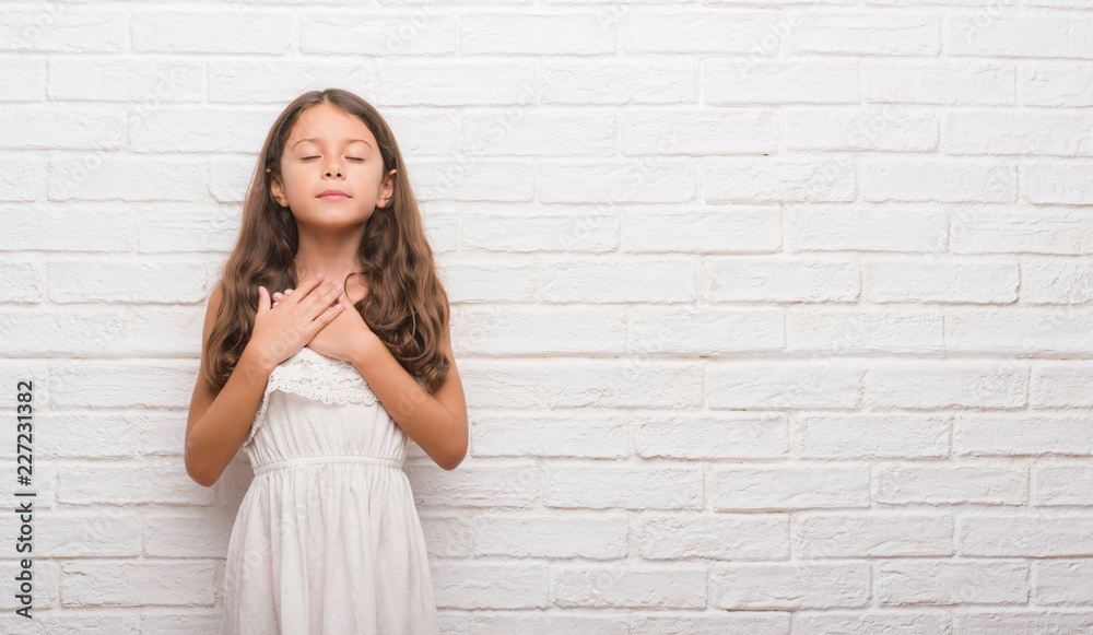 Young hispanic kid over white brick wall smiling with hands on chest with closed eyes and grateful gesture on face. Health concept.