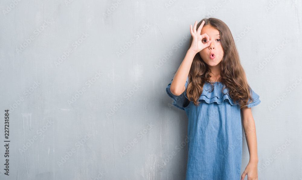 Young hispanic kid over grunge grey wall doing ok gesture shocked with surprised face, eye looking through fingers. Unbelieving expression. Young hispanic kid over grunge grey wall doing ok gesture shocked with surprised face, eye looking through fingers. Unbelieving expression.
