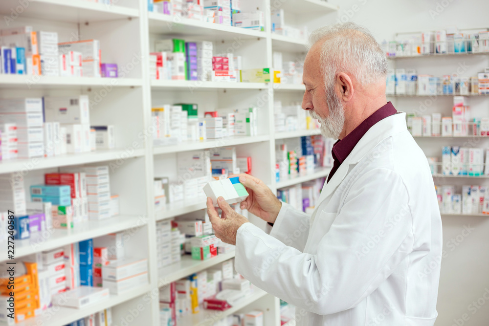 Senior male pharmacist holding a box of medications and reading labels. Side view