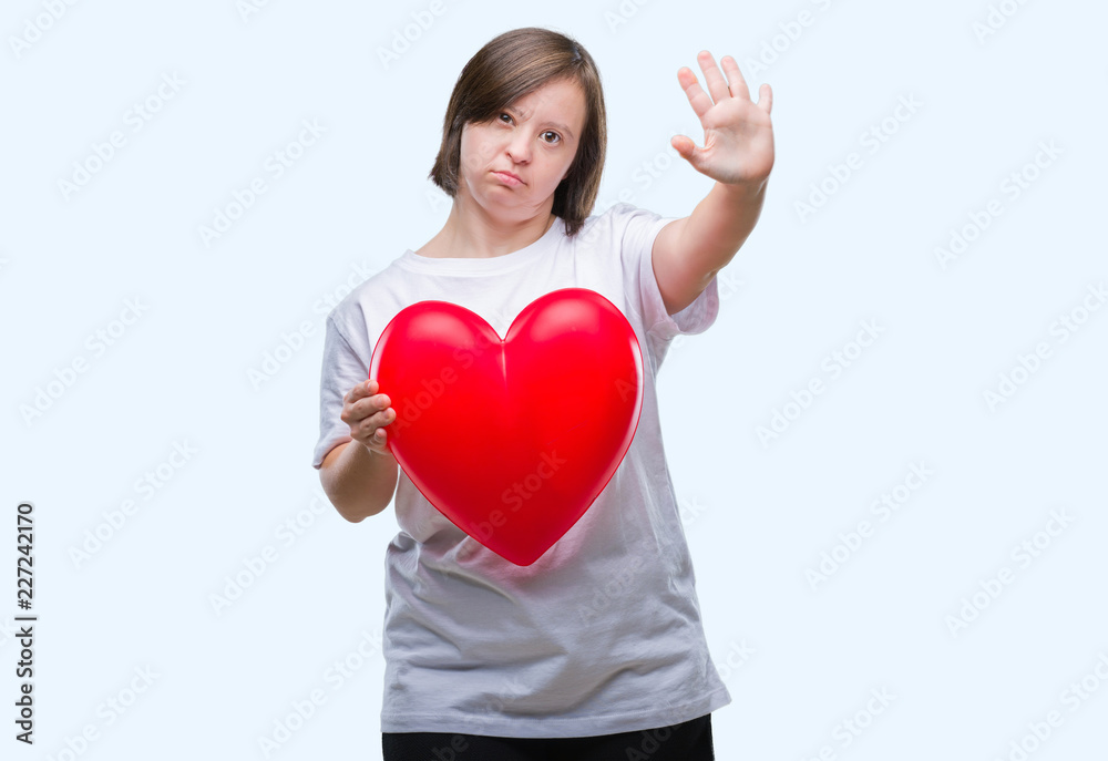Young adult woman with down syndrome holding red heart over isolated background with open hand doing stop sign with serious and confident expression, defense gesture Young adult woman with down syndrome holding red heart over isolated background with open hand doing stop sign with serious and confident expression, defense gesture