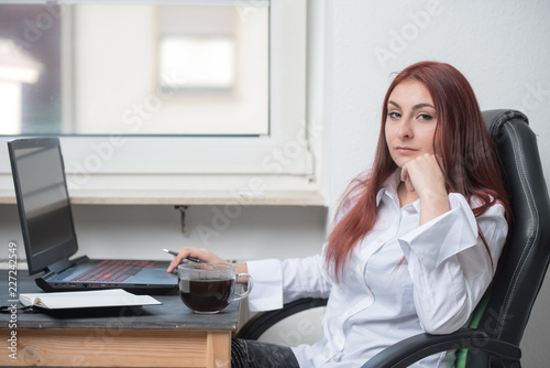 A young woman in a white shirt is sitting at a desk by the window. An open laptop, coffee and notebook are on the desk. She is looking directly at the viewer