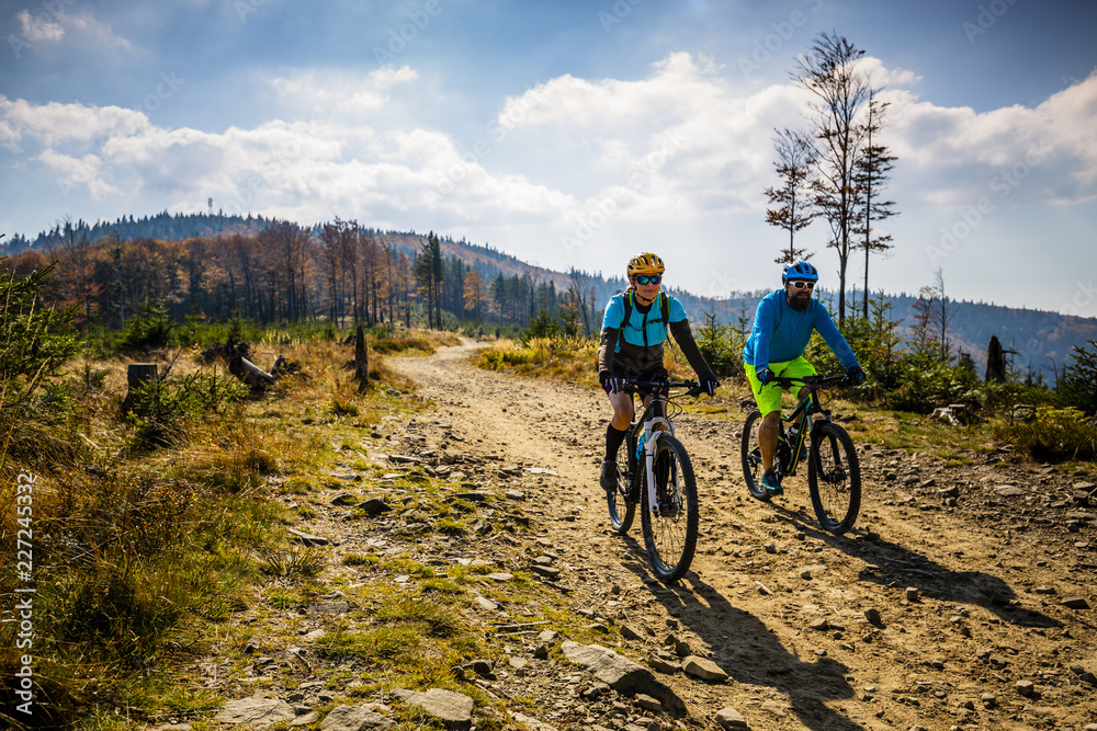 Mountain biking woman and man riding on bikes at sunset mountains forest landscape. Couple cycling MTB enduro flow trail track. Outdoor sport activity.