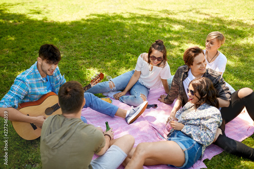 friendship, leisure and summer concept - group of happy smiling friends with guitar and non alcoholic drinks chilling on picnic blanket at summer park