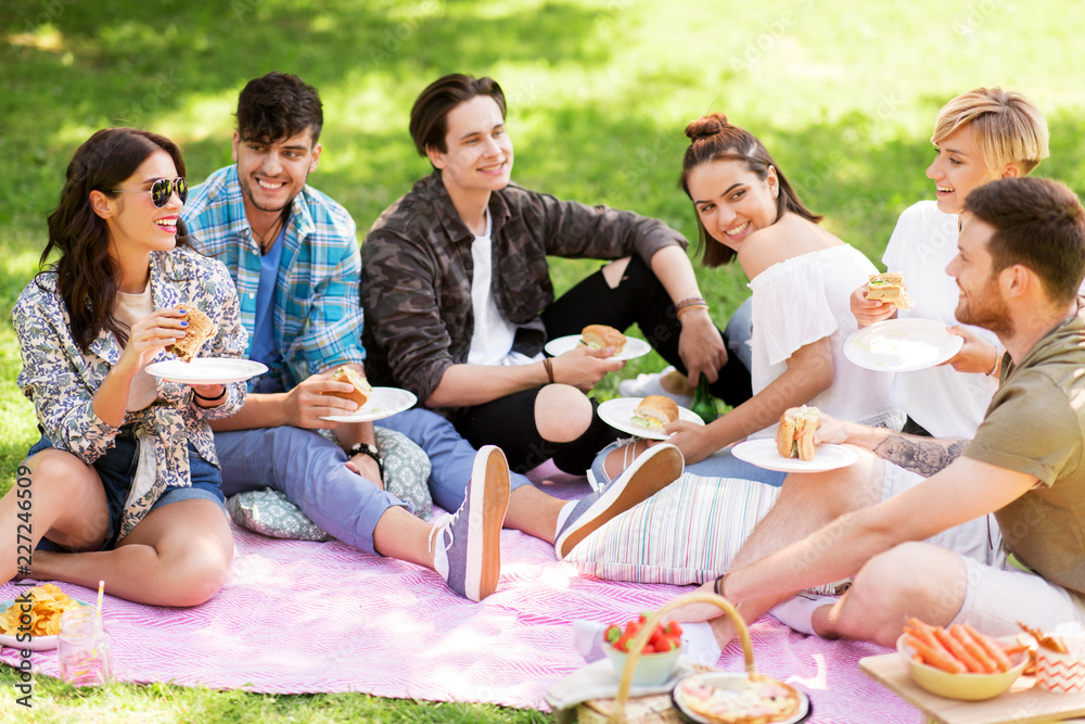 friendship, leisure and fast food concept - group of happy friends eating sandwiches or burgers at picnic in summer park