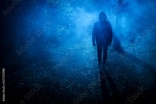 Halloween concept. Man Maniac in white mask and black hood in the forest under the blue light of the moon and fog at night, look.