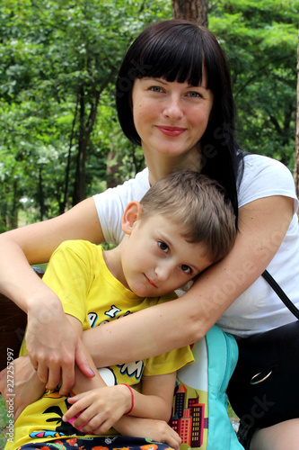 young female model smiling while looking at the camera, sitting on a bench in the park with her little son.