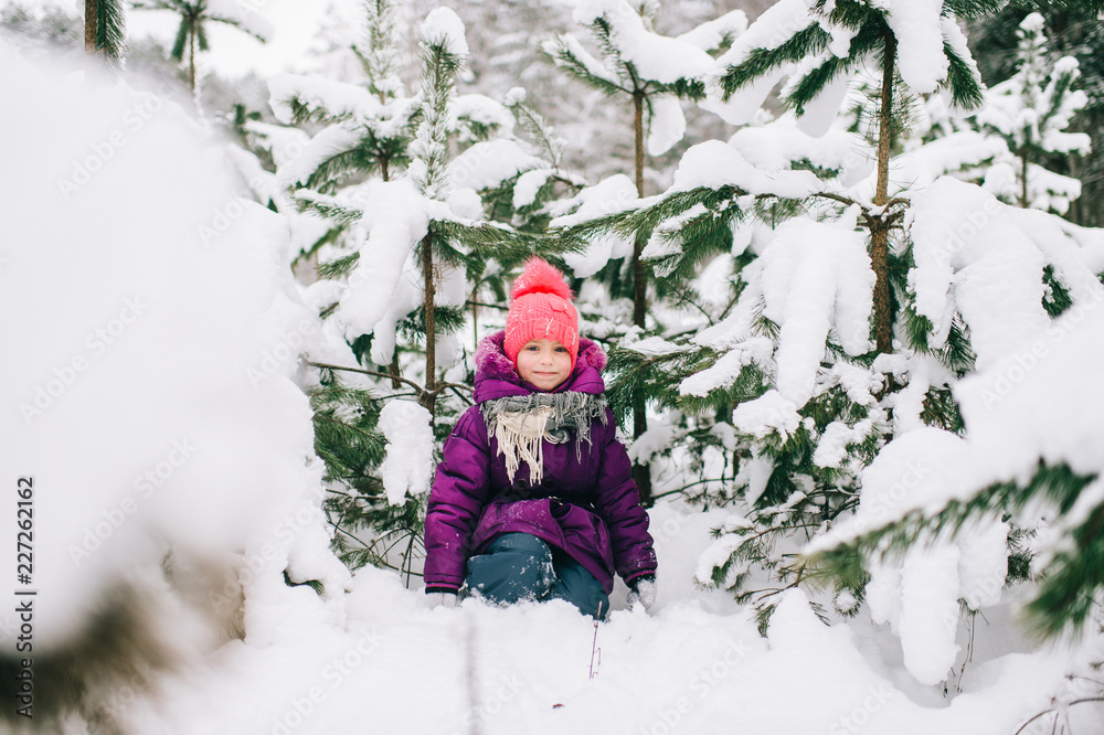 Adorable little girl posing under snowy spruce tree in wood outdoor. Beautiful female child sitting on snow in front of christmas tree in forest. Winter portrait of lovely young baby. Funny kid face. Adorable little girl posing under snowy spruce tree in wood outdoor. Beautiful female child sitting on snow in front of christmas tree in forest. Winter portrait of lovely young baby. Funny kid face.