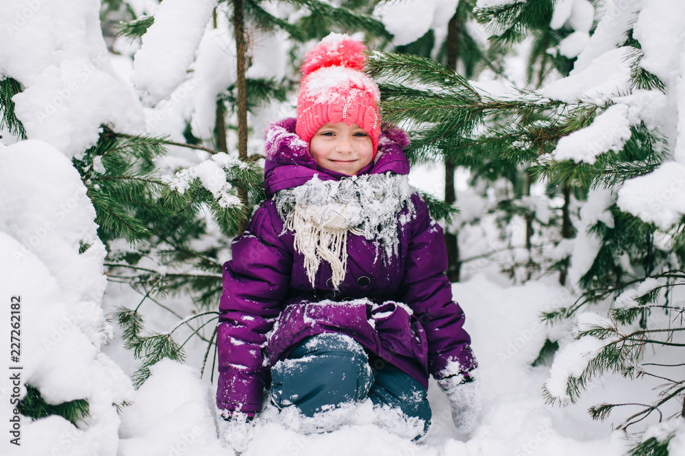 Adorable little girl posing under snowy spruce tree in wood outdoor. Beautiful female child sitting on snow in front of christmas tree in forest. Winter portrait of lovely young baby.  Funny kid face.