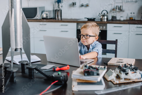 funny adorable boy using laptop at table with rocket model in kitchen on weekend