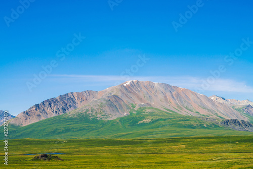 Giant mountains with snow above green valley under clear blue sky. Meadow with rich vegetation and lakes of highlands in sunlight. Amazing sunny mountain landscape of majestic nature.