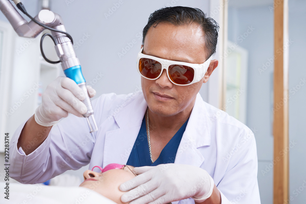 Male specialist in protective glasses lifting laser rejuvenation device under female face at beauty salon Male specialist in protective glasses lifting laser rejuvenation device under female face at beauty salon