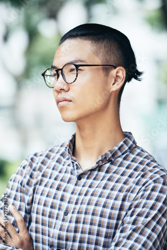 Waist-up portrait of young Asian man in checked shirt and glasses standing outdoors and looking into the distance on blurred background