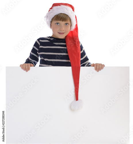 Cute boy wearing Santa Claus hat, posing behind white panel isolated on white background. Child holding empty Christmas billboard. Boy peeping behind blank holidays board. photo