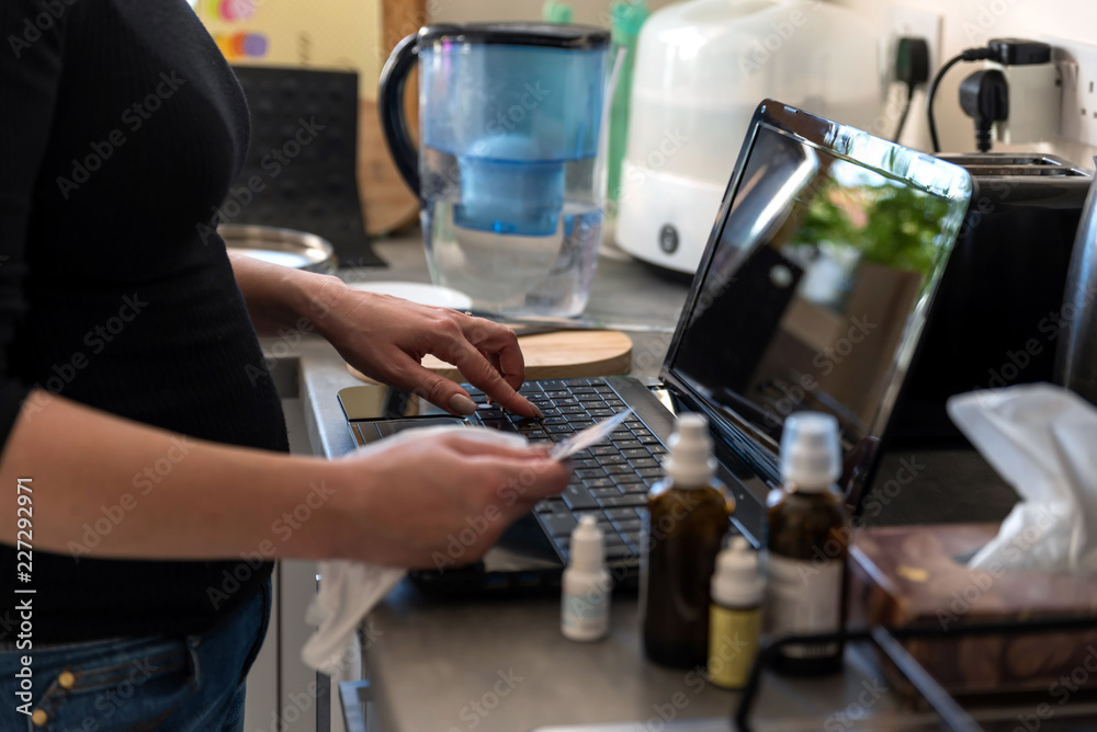 young sick woman at home kitchen paying for mdeicines on laptop next to medicines