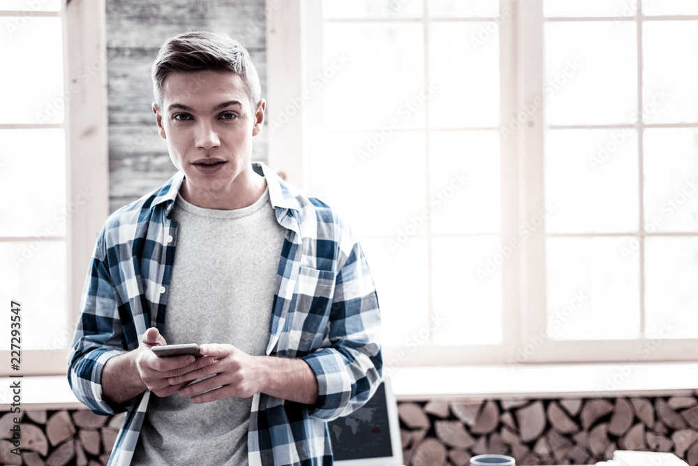 Handsome person. Calm handsome student being at home and looking relaxed while standing with a modern smartphone in his hands Handsome person. Calm handsome student being at home and looking relaxed while standing with a modern smartphone in his hands