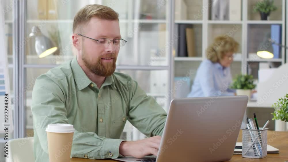 Young Caucasian businessman in eyeglasses sitting at desk in the office, writing on laptop and smiling at camera Young Caucasian businessman in eyeglasses sitting at desk in the office, writing on laptop and smiling at camera