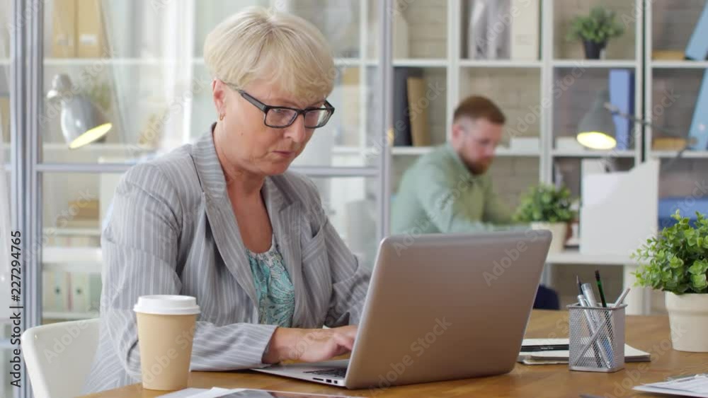 Concentrated senior businesswoman in eyeglasses sitting at desk in the office and typing on laptop Concentrated senior businesswoman in eyeglasses sitting at desk in the office and typing on laptop