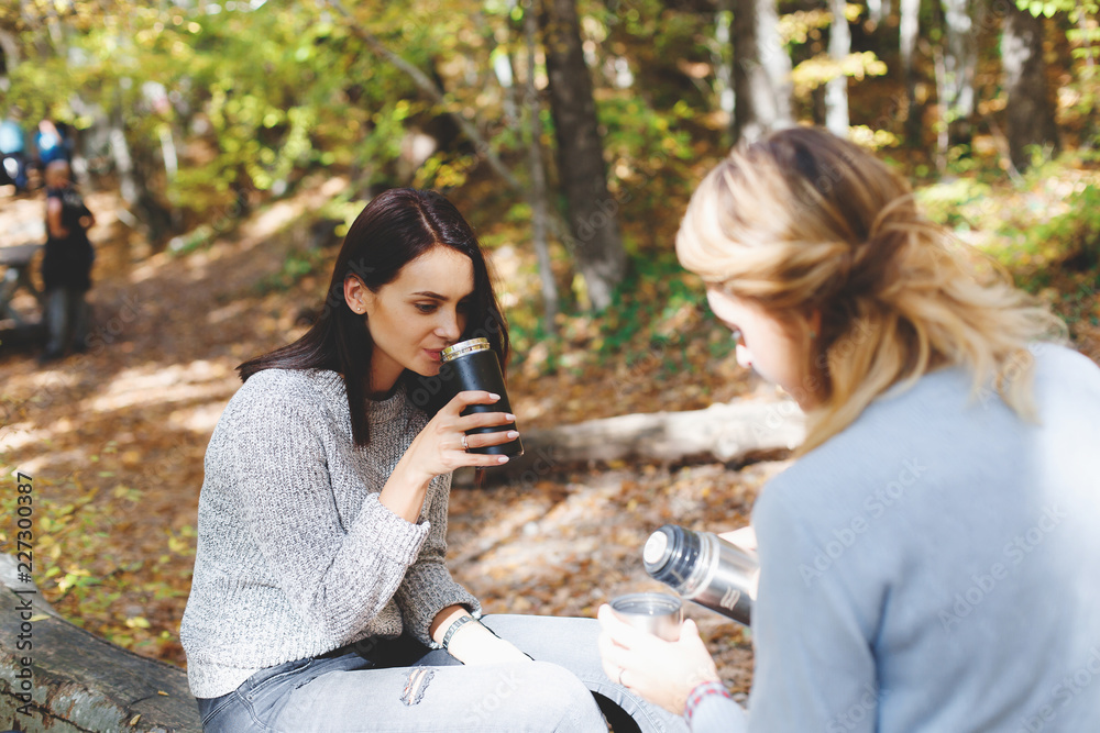 Two young girls girlfriend sat down to rest and drink tea from thermo cup in the autumn forest. Walk in the woods. Stop for a rest and a snack. Girlfriends hiking in the fall. Two young girls girlfriend sat down to rest and drink tea from thermo cup in the autumn forest. Walk in the woods. Stop for a rest and a snack. Girlfriends hiking in the fall.
