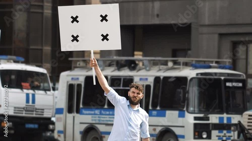 One bearded european guy on the background of a police car holds a banner in hand and look at camera. Protest 1 caucasian man on street in city. Angry one human picketing on background of bus cops 4k. photo