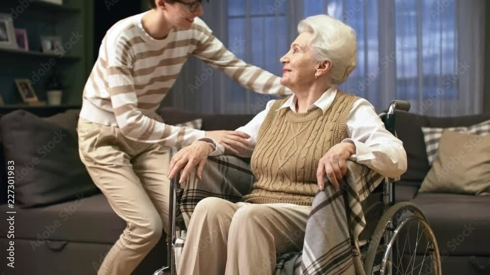 Tilt up of happy young woman and her senior grandmother in wheelchair looking at camera and posing
