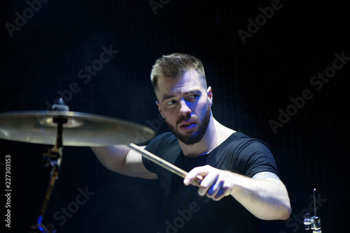 Drummer in a cap and headphones plays drums at a concert under white light in a smoke