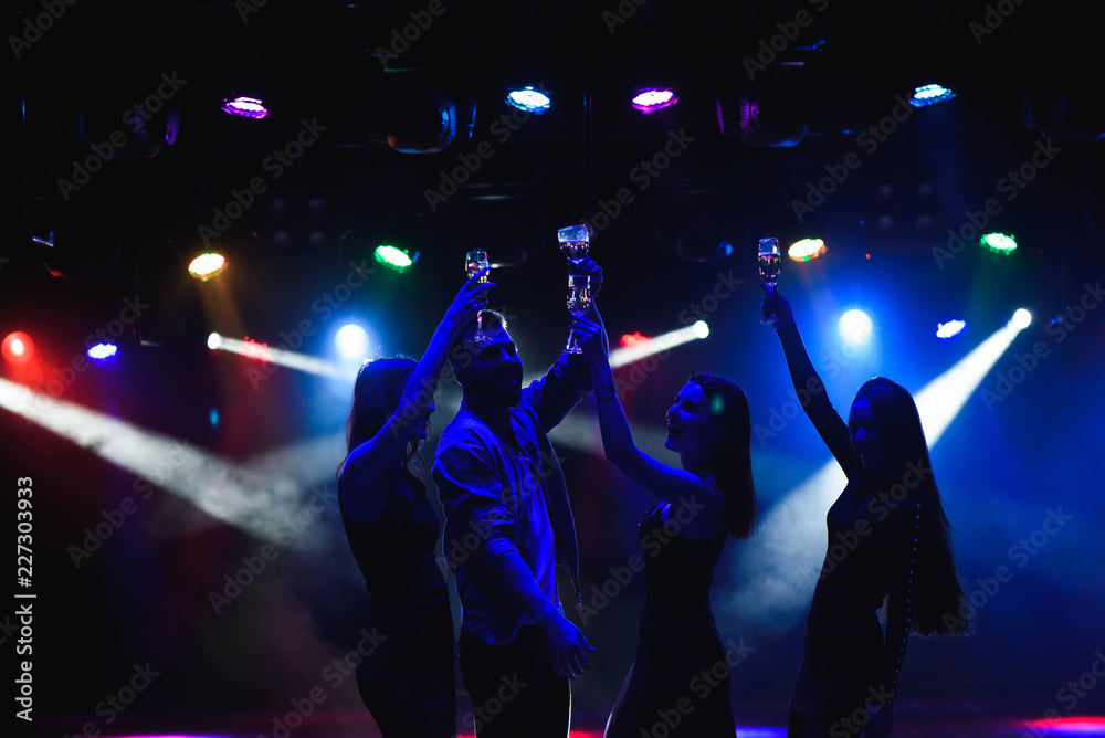 Fototapeta premium Young friends dancing with glasses of champagne in hands. Against lighting devices as background. Young people's friends are dancing.
