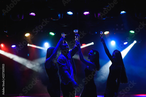  Young friends dancing with glasses of champagne in hands. Against lighting devices as background. Young people's friends are dancing.