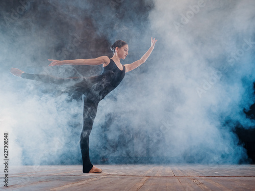 Young beautiful ballerina on smoke stage dancing modern ballet. performs smooth movements with hands against spotlights background. Woman in black costume on scene © kohanova1991