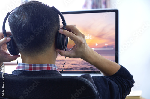 Back side of an Asian man wearing black heaphones in front of desktop computer screen listen to a music streaming with entertainment music application screen. Copy space included.