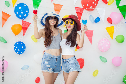 two cheerful asian young woman in big summer hat dance and drinking champagne and alcohol together in party on balloons and flag decoration background . Happy Girl celebrate in holiday