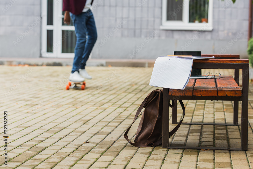cropped shot of student riding skateboard with backpack and notebook on bench on foreground