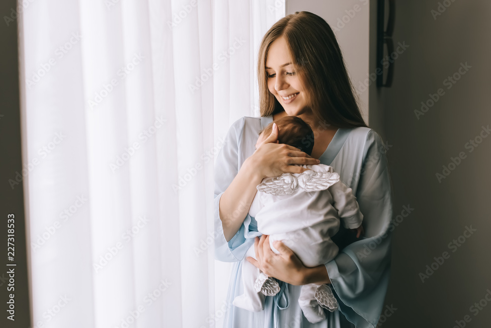 smiling attractive woman carrying little baby boy in front of curtains - obrazy, fototapety, plakaty 