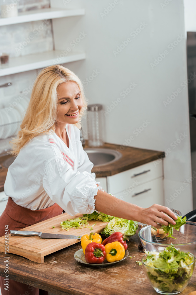 attractive woman preparing salad for dinner and putting ingredients in bowl at home