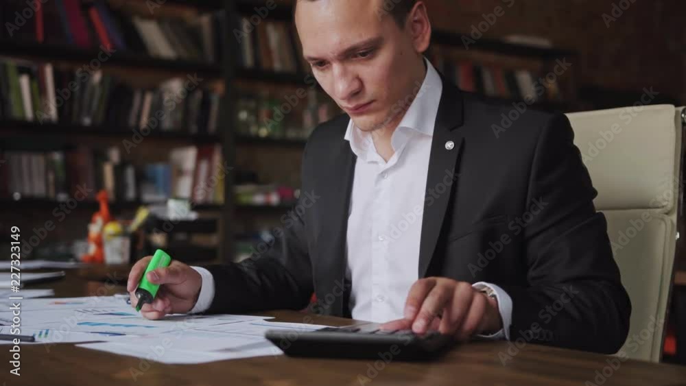 Young businessman wearing suit working with documents and calculator sitting at office desk, financial analyst using or economist analyzing statistics. Business, people, paperwork concept Young businessman wearing suit working with documents and calculator sitting at office desk, financial analyst using or economist analyzing statistics. Business, people, paperwork concept