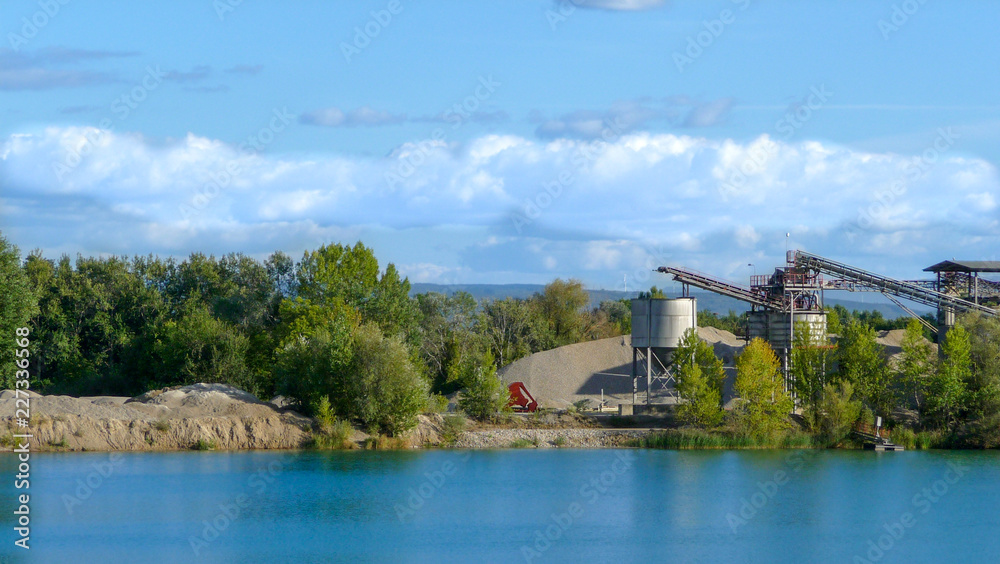 Landscape with a bright blue lake, green trees, beautiful clouds and structures for the extraction of pebbles.