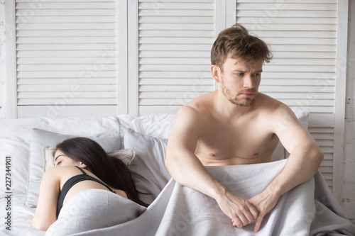 Young millennial attractive couple lying in bed under blanket at home. Woman sleeping, depressed and frustrated man thinking about relationships. Family have problems in sexual life impotence concept photo