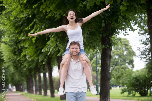 Young attractive couple in love wear casual summer clothes spend free time together in park. Happy girl sits on shoulders of beloved guy. Romantic relations or friendship between man and woman concept