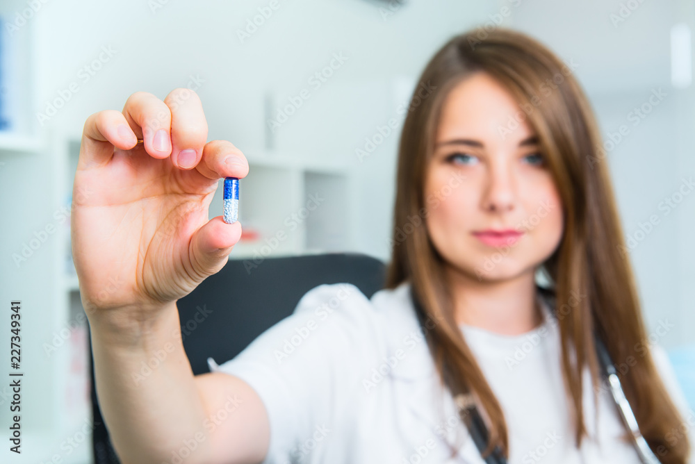 Smiling Female doctor in white coat with stethoscope holding a pill between her fingers. Hand of a hospital medical expert shows the pill to be taken to patient. Healthcare and medical concept.